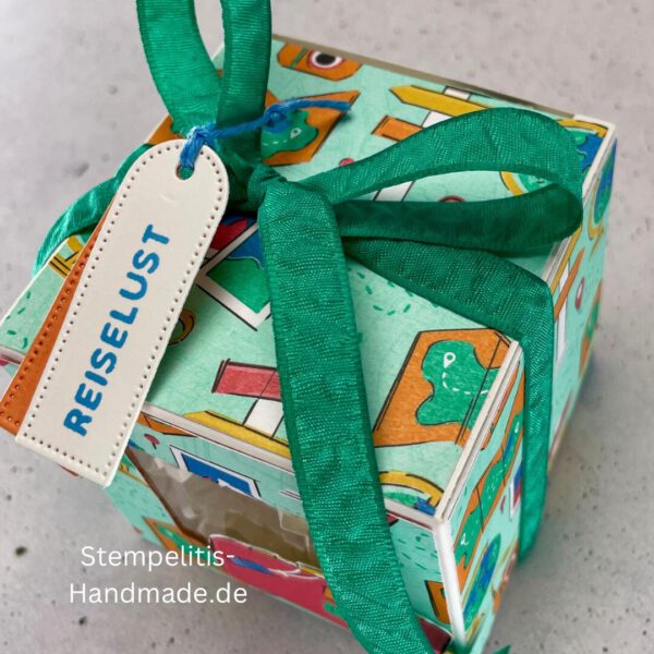 Handdekorierte Fensterschachtel mit Reisethema und grünem Band, Detailansicht des Sichtfensters mit Füllmaterial, Koffer-Motiv auf der Front der Schachtel, Oberseite mit Geschenkanhänger „Reiselust“, Seitliche Ansicht der Box mit Weltkartenmotiv, Geöffnete Box mit Blick auf das Innere, Fensterschachtel als kreative Geschenkverpackung für Reisegutscheine